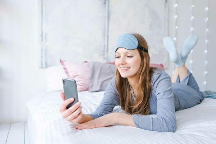 Happy young woman relaxing using and talking with smartphone on bed at home